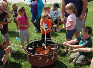 Summer reading program participants gathered around the fire they made to cook bannock bread on sticks.  Pictured here, from left to right, are Kayleigh Cole, Isabella Cole, Cienna Bishop, Owen Sheltra, Brielle Rancourt, and Trevor Sanville.  Directly behind Owen are Chase Sheltra who is looking at his dough-covered stick, and Dale Guisinger, who is digging into a Tupperware for more dough to hand out.  Photo by Nathalie Gagnon-Joseph