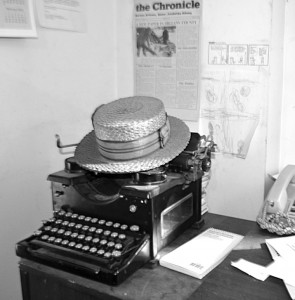 A solid reminder of how we used to operate — an old manual typewriter — sits in a corner of the Chronicle office.  The hat belonged to Anna Baker, the artist responsible for the Chronicle cows, and on the wall behind it is a copy of the original flyer announcing the start of a new newspaper, the Chronicle.  Photo by Bethany M. Dunbar